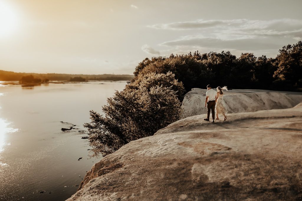 Engaged couple overlooking the Susquehanna River from atop the White Cliffs of Canoy during a golden hour sunset in a green dress and brown pants